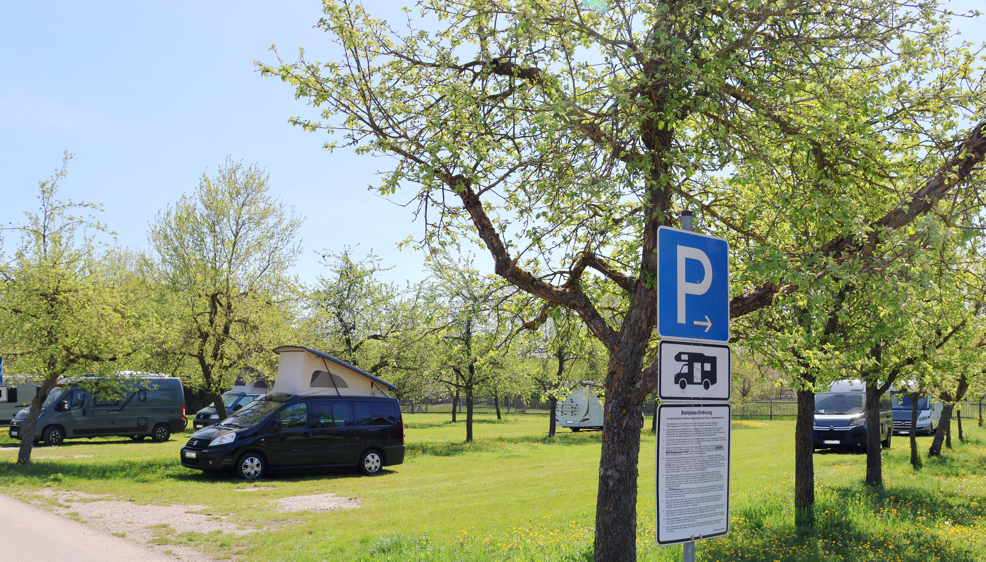 Grüne Wiese mit Bäumen im Frühjahr. Im Vordergrund ein blaues Schild mit einem Parken für Wohnmobile Zeichen. Im Hintergrund Wohnmobild.