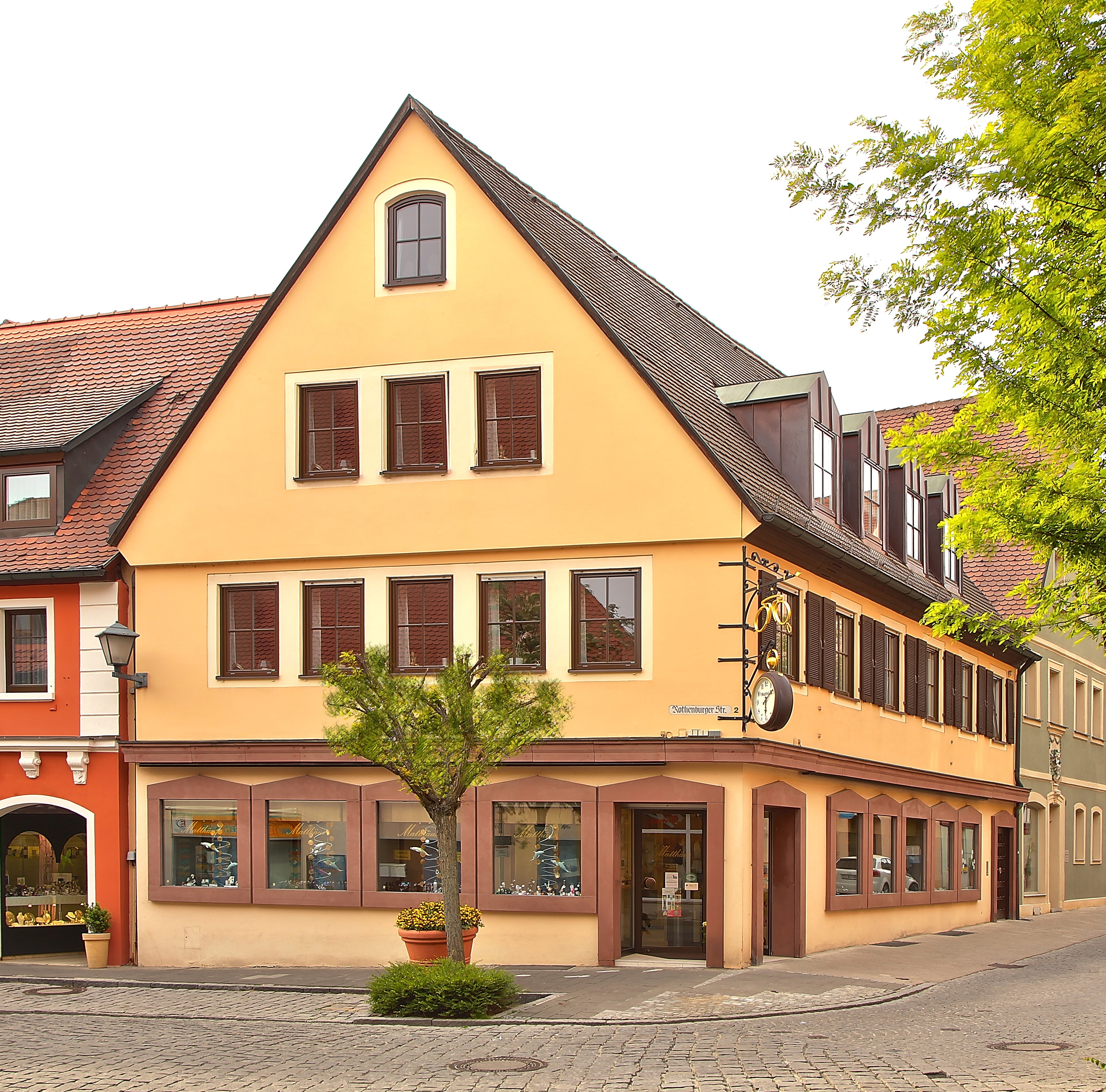 Ein Eckgebäude in der Innenstadt. Dreistöckig, gelb unten ein Geschäft. Vor dem Haus ein kleiner Baum