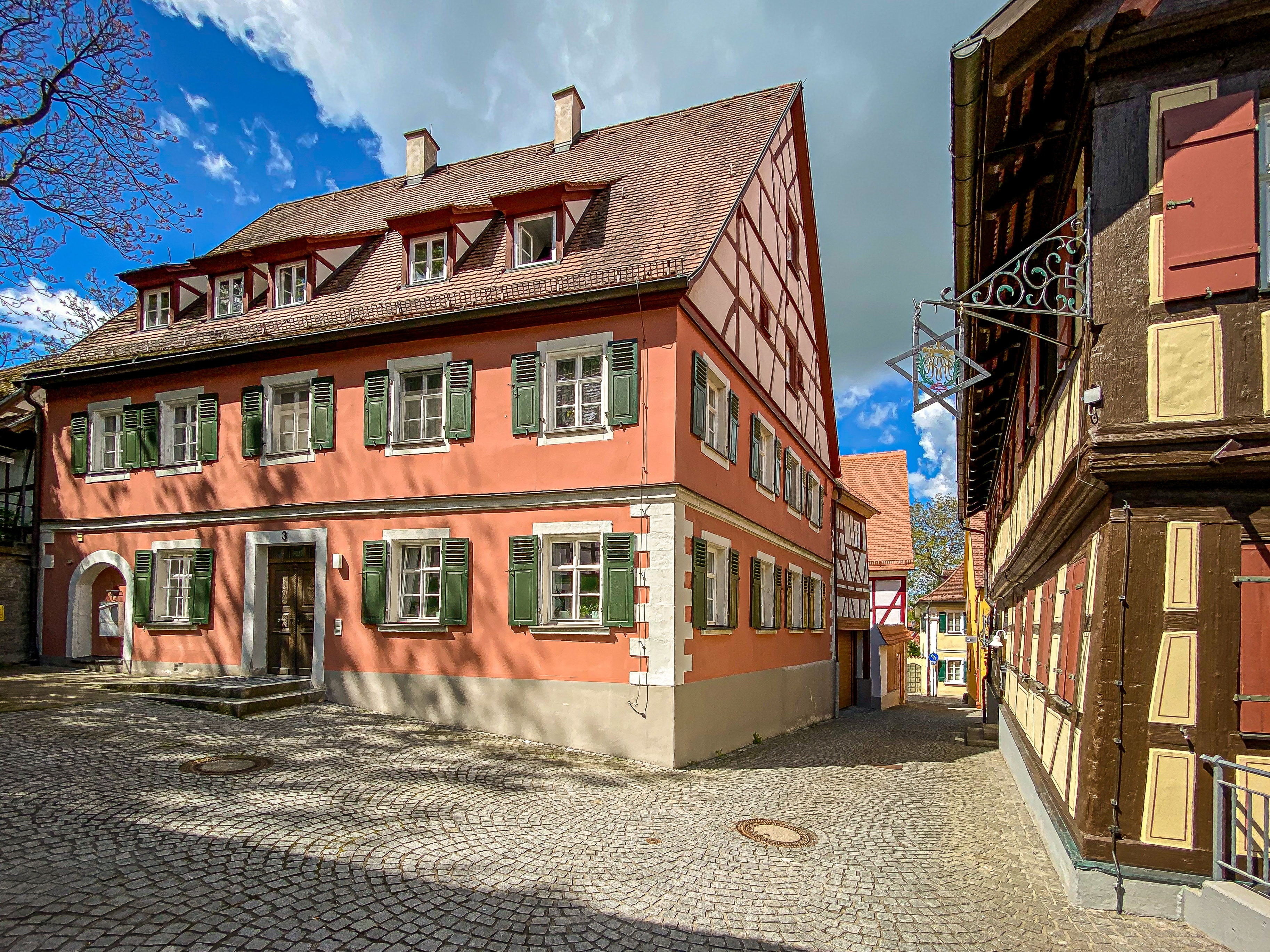 Ausschnitt einer Altstadtgasse in Bad Windsheim, korallenfarbenes Steinhaus mit grünen Holzfensterläden und Fachwerkgiebel, rechts im Bild angeschnittene Traufseite eines historischen Fachwerkhauses mit einem Metallschild der Brauerzunft, Kopsteinpflaster