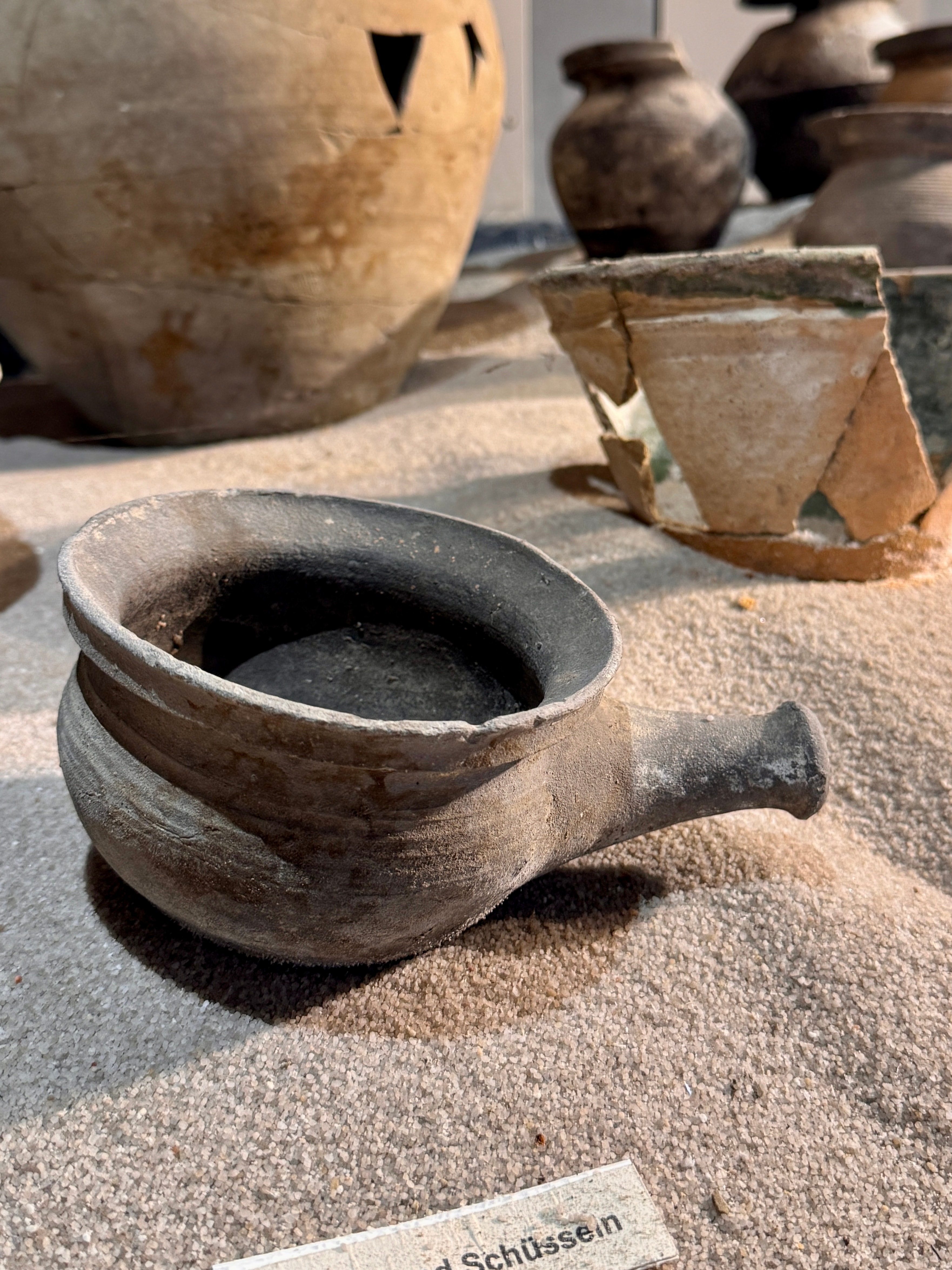 Scherben und ein Steinguttop aus der Ausstellung im Archäologischen Fenster der Stadt Bad Windsheim. Die Ausstellungsstücke sind in Sand gebettet und beleuchtet