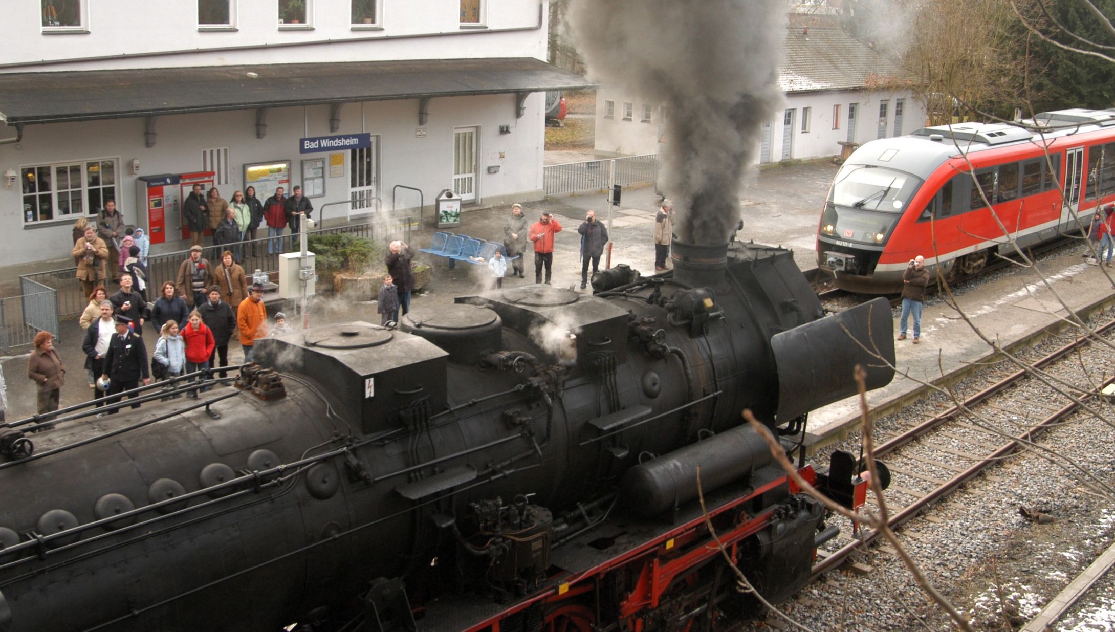 Alte Dampflok, die grauen Rauch ausstößt, im Bad Windsheimer Bahnhof mit einfahrender moderner Diesel-Regionalbahn; Menschenauflauf, der die Dampflok bestaunt und fotografiert