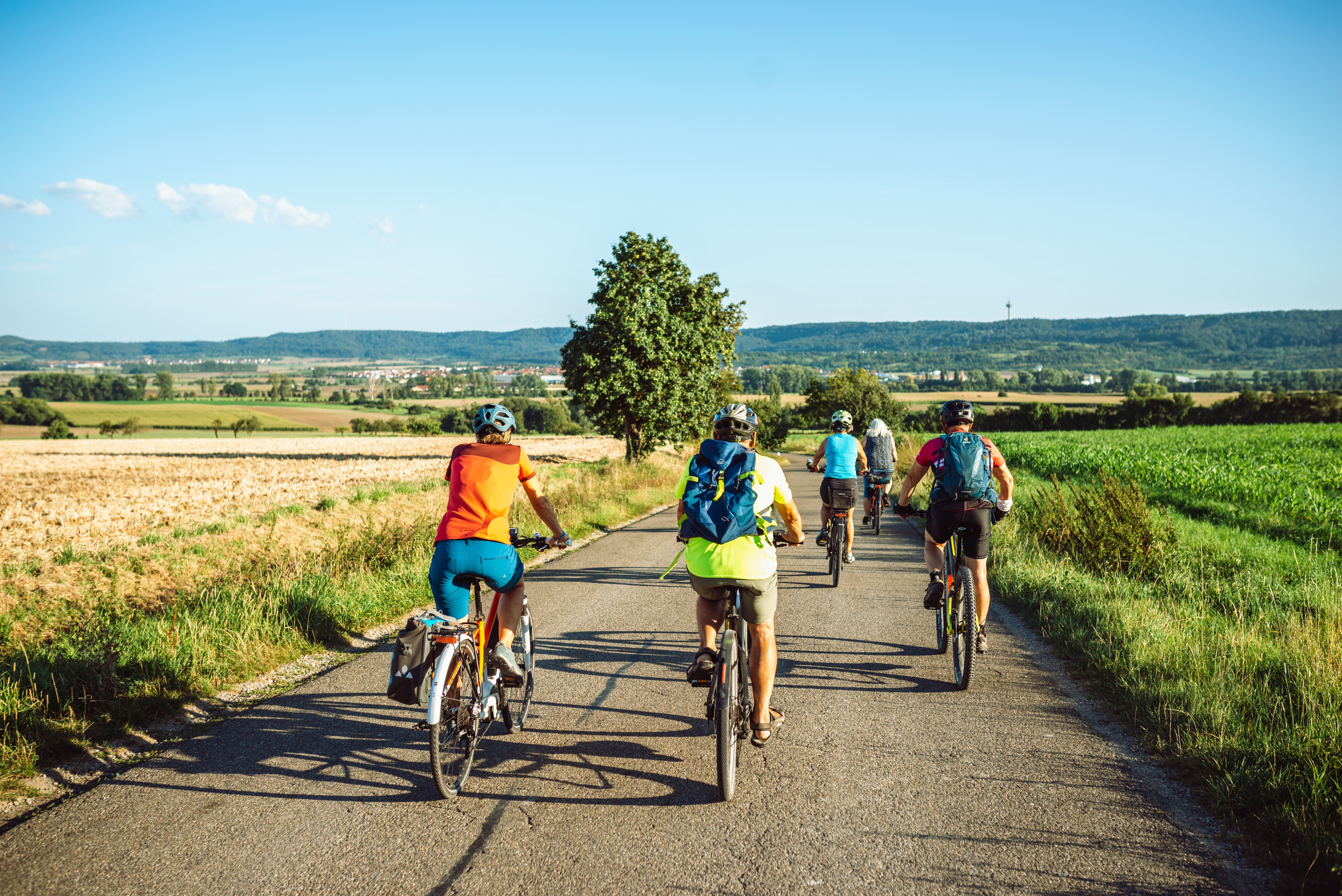 Eine Gruppe Radfahrender radelt auf einem Wirtschaftsweg durch gelbgolde Getreidefelder und grüne Wiesen, am Horizont ist sind die Ausläufer des Steigerwaldes und Burgbernheim zu sehen; die radelnden tragen sommerliche Kleidung