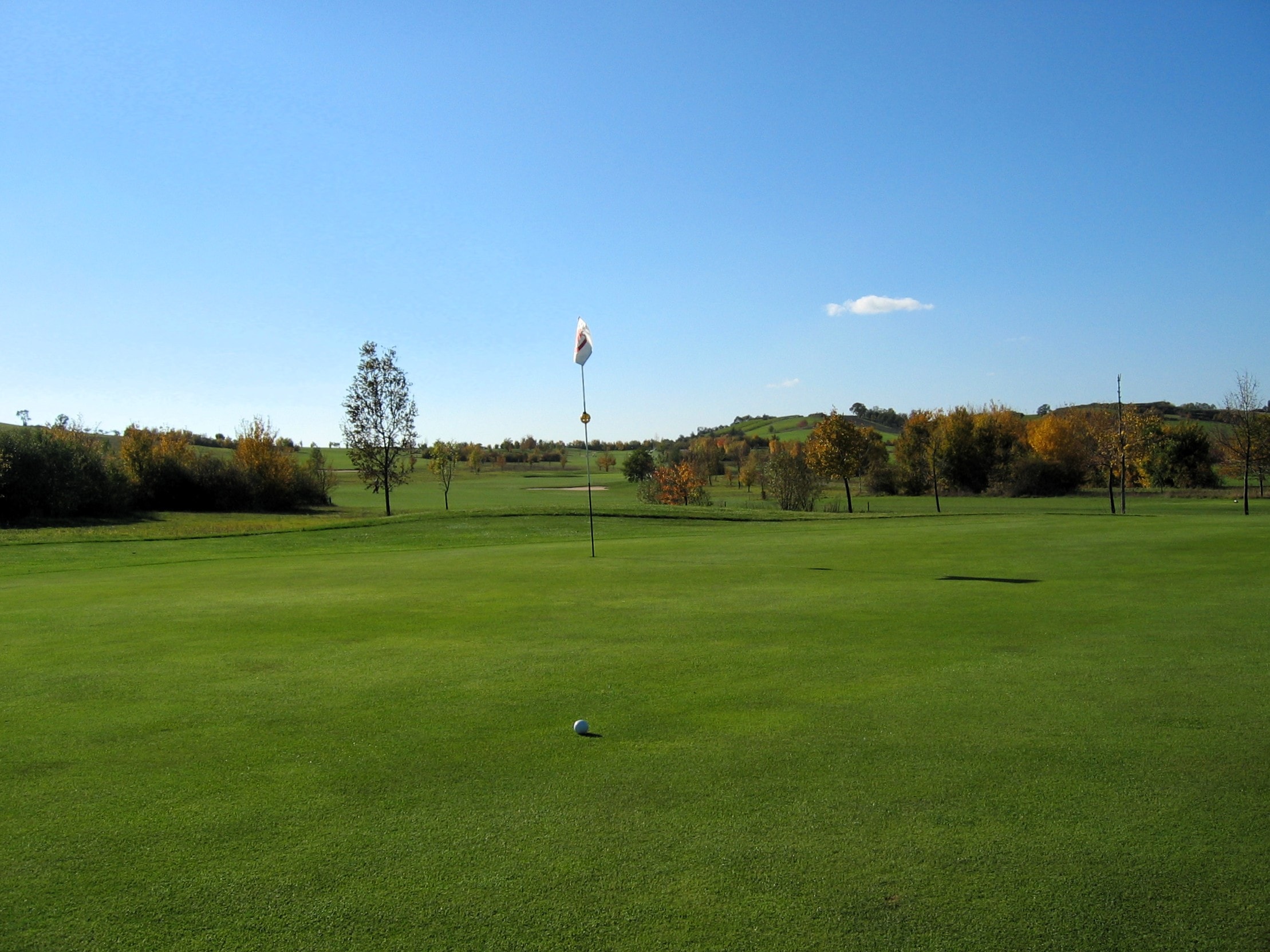 Golfplatz Bad Windsheim, Übergang Green zum Fairway, Fähnchen, Sträucher & Bäume vor blauem Himmel