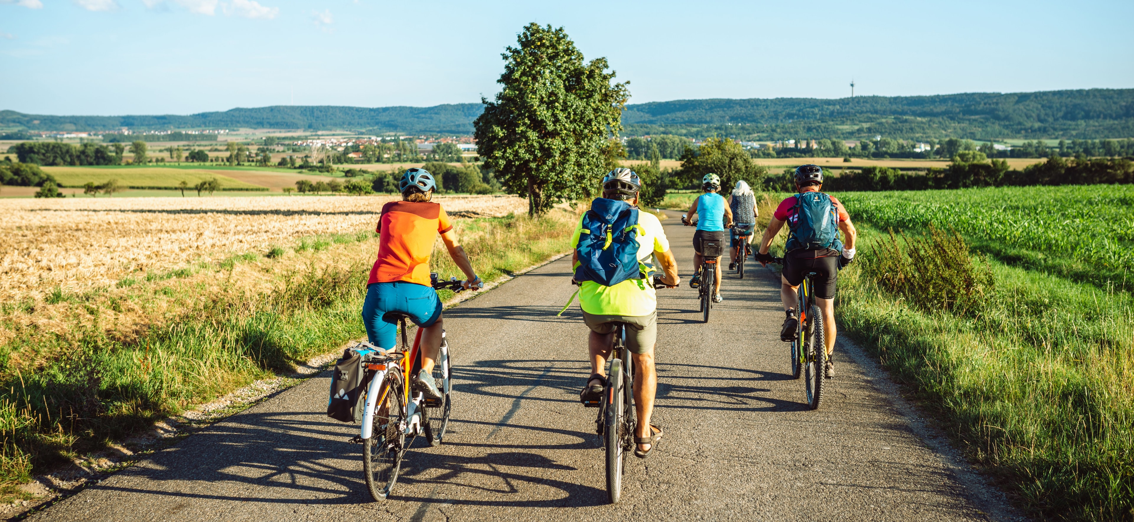 Eine Gruppe sommerlich gekleideter Radfahrender befährt einen von Feldern gesäumten Wirtschaftsweg Richtung Burgbernheim, am Horizont der Steigerwald, blauer Himmel