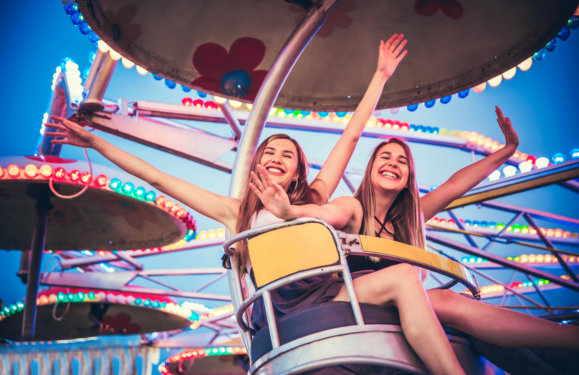 Zwei junge, sommerlich gekleidete Frauen sitzen in einer bunt beleuchteten Gondel eines Fahrgeschäfts, lachen und winken mit erhobenen Armen, Hintergrund blauer Himmel