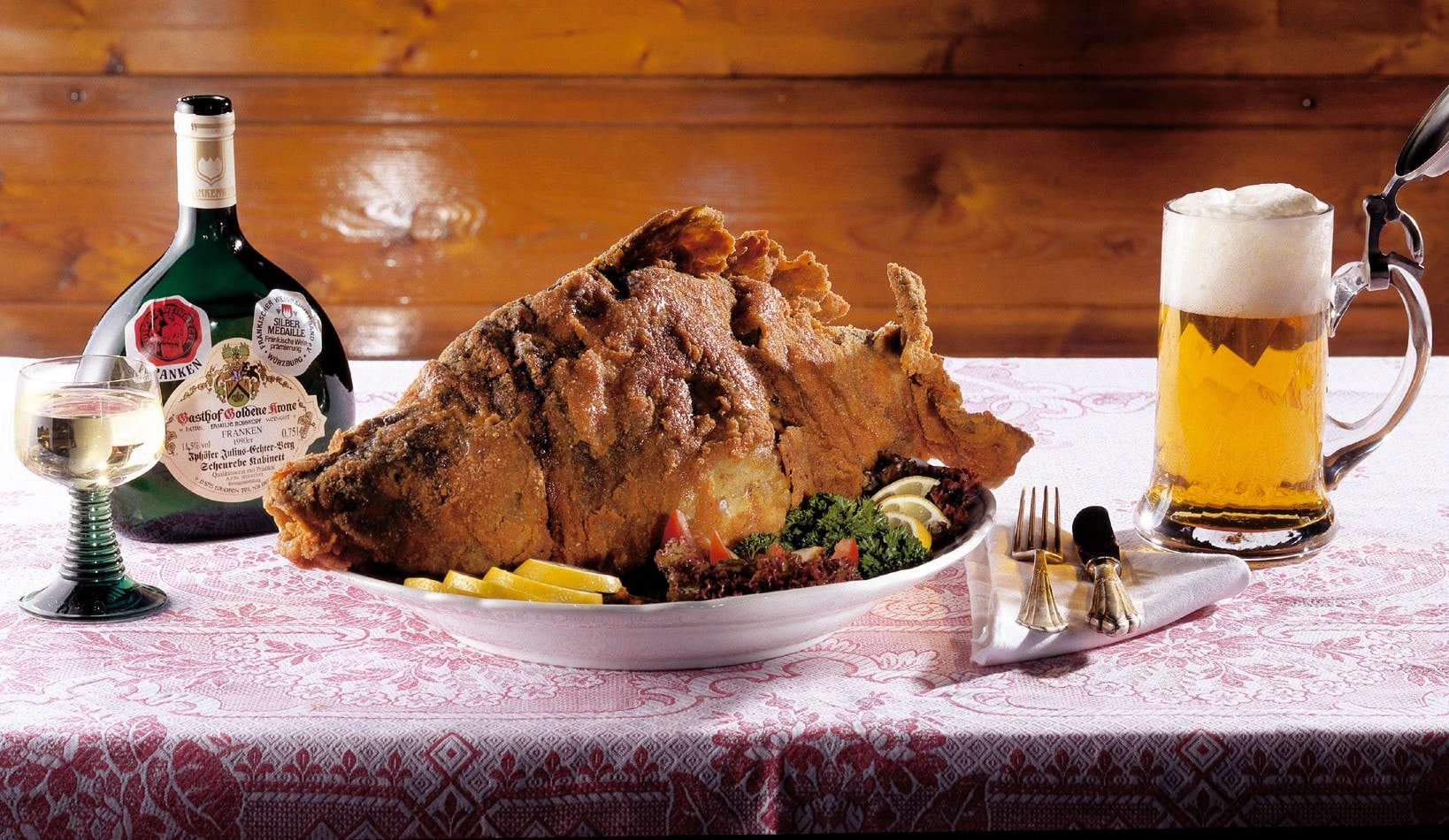 Aischgründer Karpfen gebacken in einer weißen Schale auf einer rot bestickten Leinentischdecke, Silberbesteck, links ein Bocksbeutel mit Römer, rechts ein frisch gezapftes Bier im Glas, im Hintergrund Holzvertäfelung