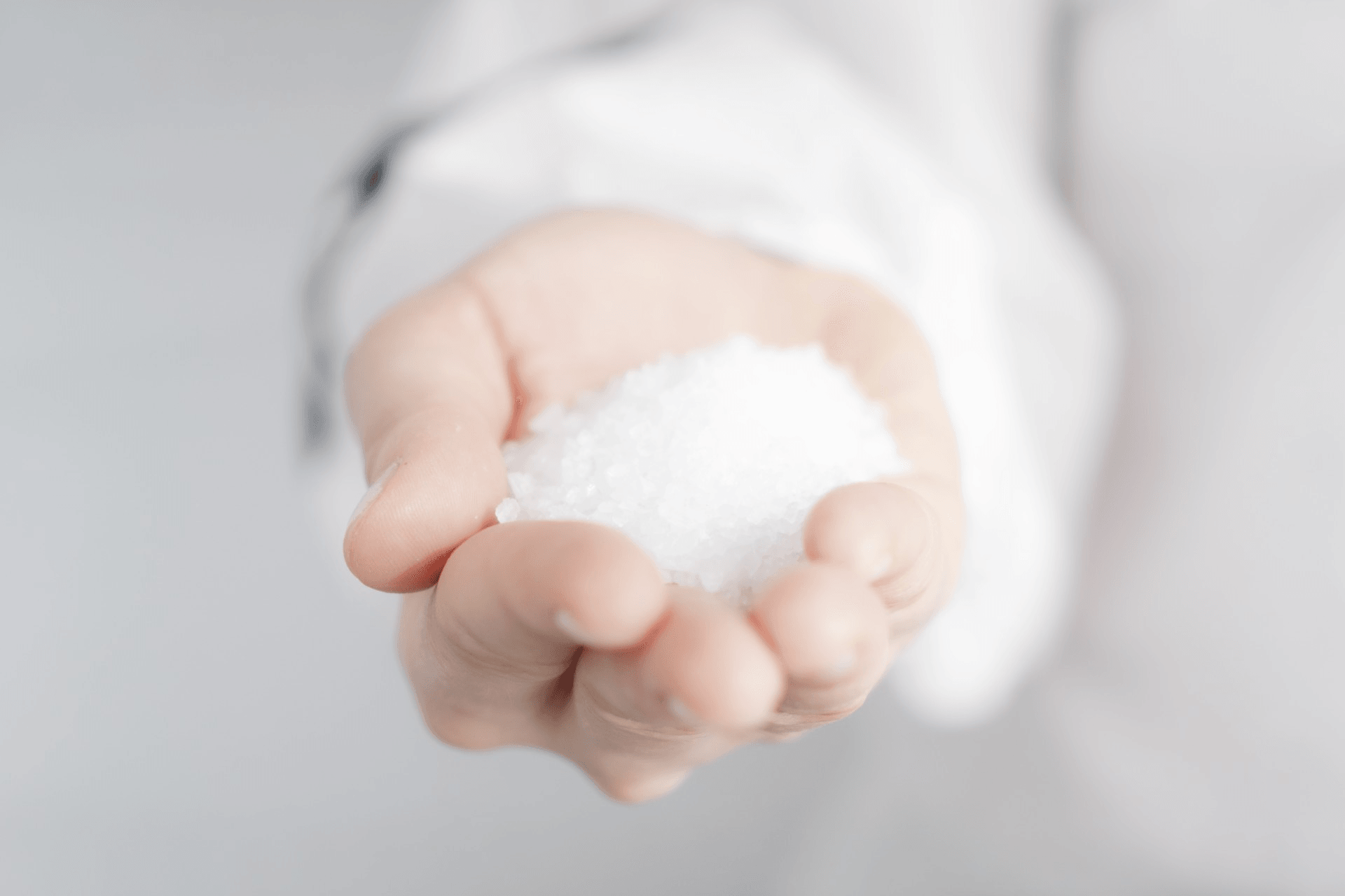 Grauer, unscharfer Hintergrund, eine Hand mit Salz befüllt streckt sich dem Betrachter entgegen. 