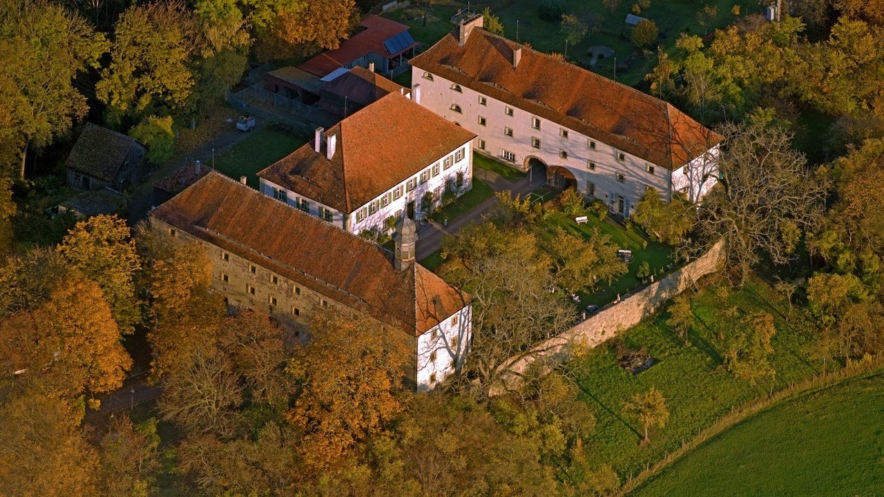 Ein altes Schloss aus drei Gebäuden in U-Form umgeben von Bäumen. Aufnahme von oben 