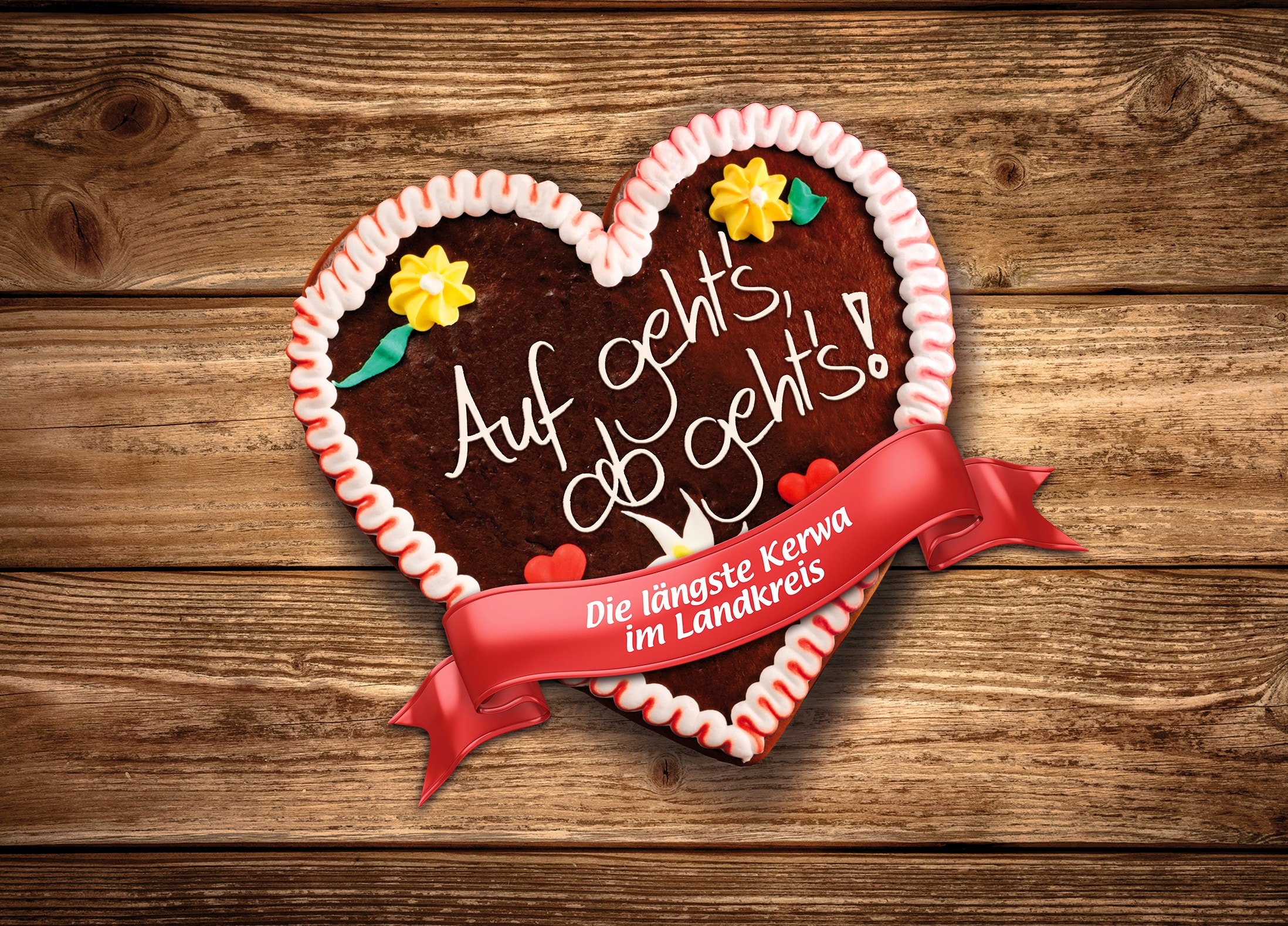 Ein Lebkuchenherz mit rot-weißem Zuckerrand, gelben Blümchen und der Aufschrift "Auf geht's - Ab geht's" liegt auf einem Holzbrett