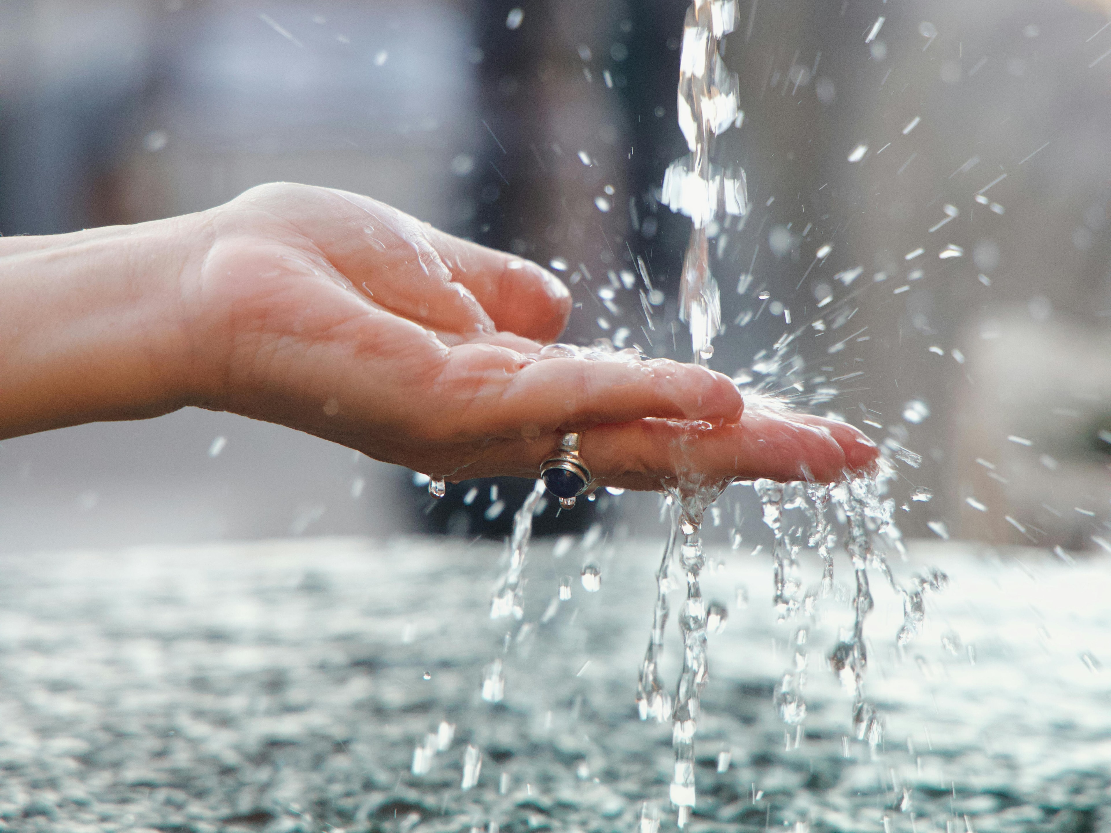 Eine Hand streckt sich unter den Quellstrahl eines Heilwasserbrunnens. Tropfen spritzen, Hintergrund unscharf.