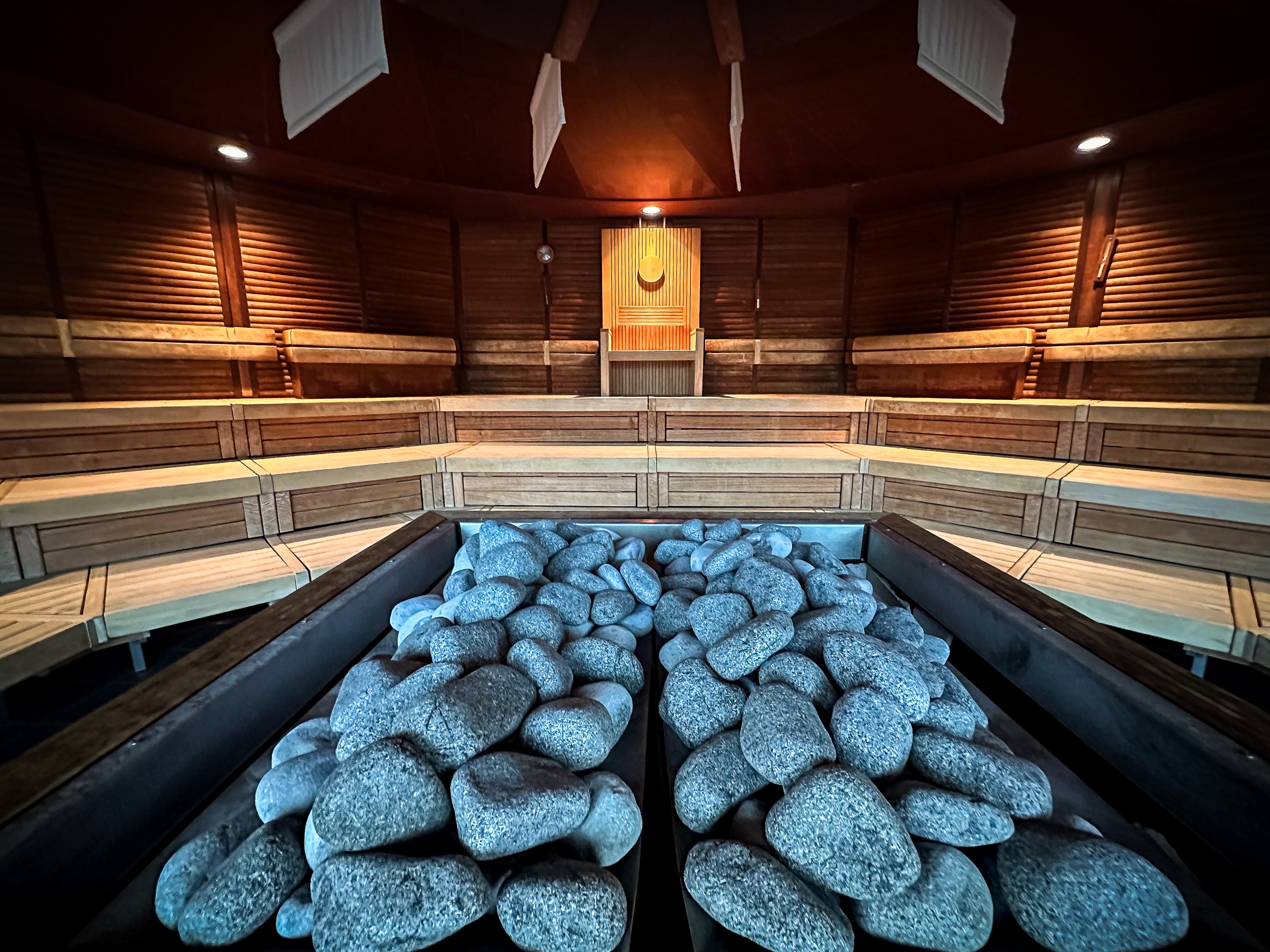 Innenansicht einer Sauna, blauschwarze Saunasteine im Vordergrund, dahinter die Stufen und ein runder Fächer am Saunahimmel