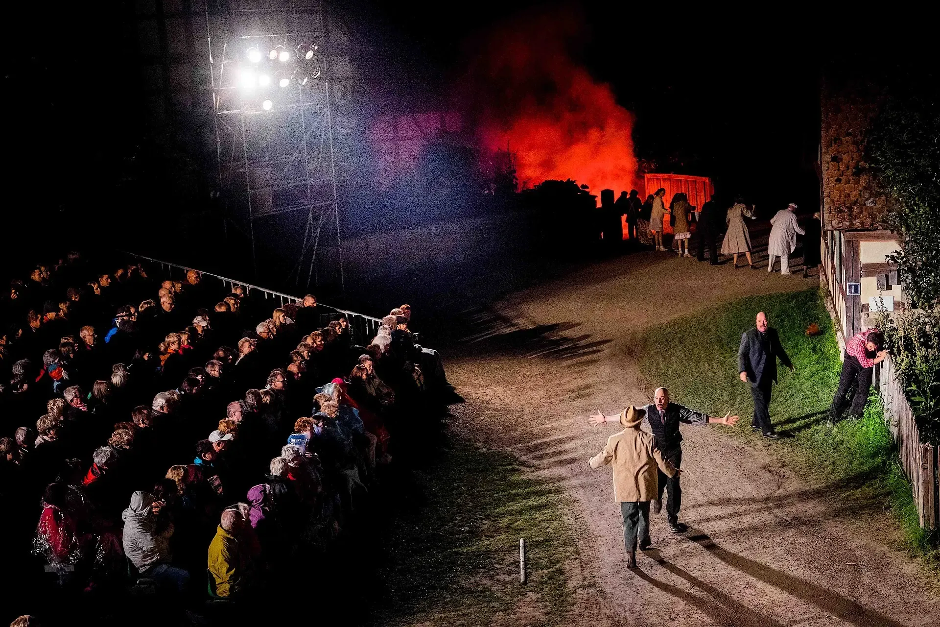 Theateraufführung, links Tribüne mit Zuschauern in der Mitte zwei Personen im Hintergrund Rauch und Feuer